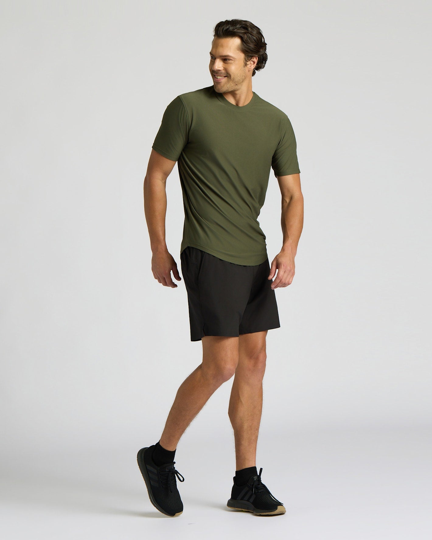 A man modeling an olive green short-sleeve athletic t-shirt and black athletic shorts against a light gray studio background.