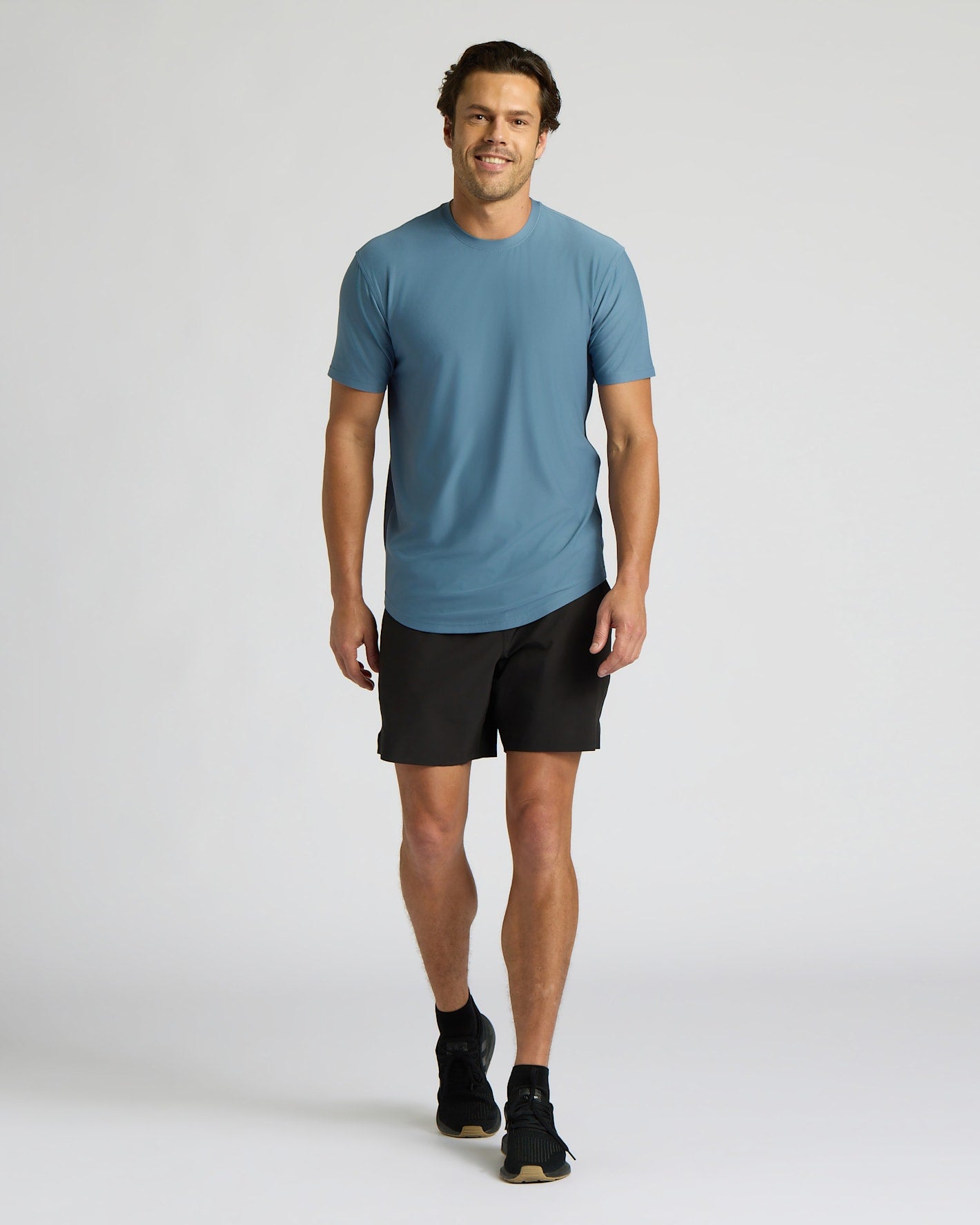 A man modeling a slate blue crewneck t-shirt and black athletic shorts against a light gray studio background.