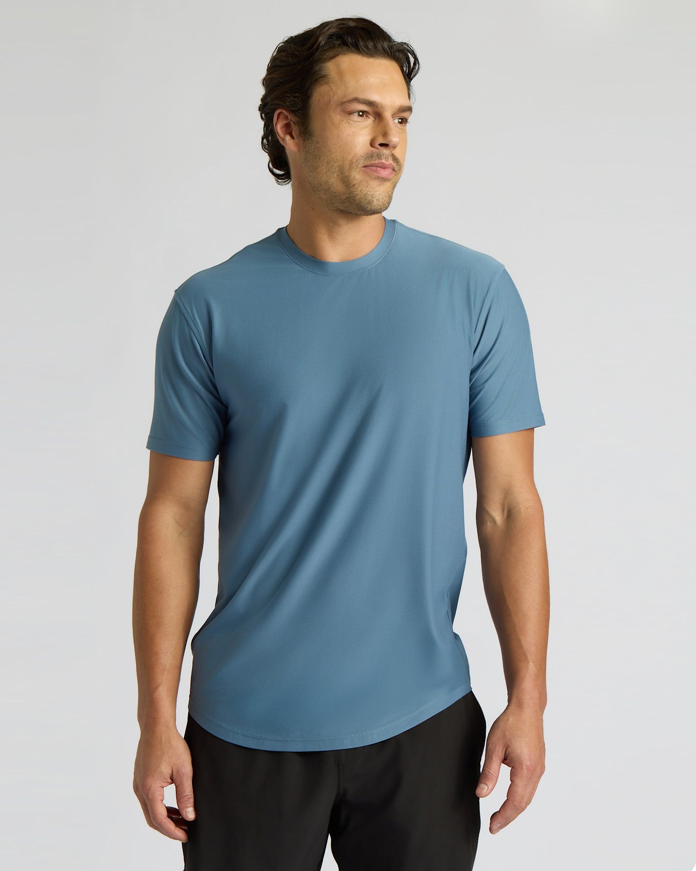 A man wearing a short-sleeve crew-neck athletic t-shirt in a muted slate blue color, paired with black athletic pants, photographed against a neutral light gray studio background.