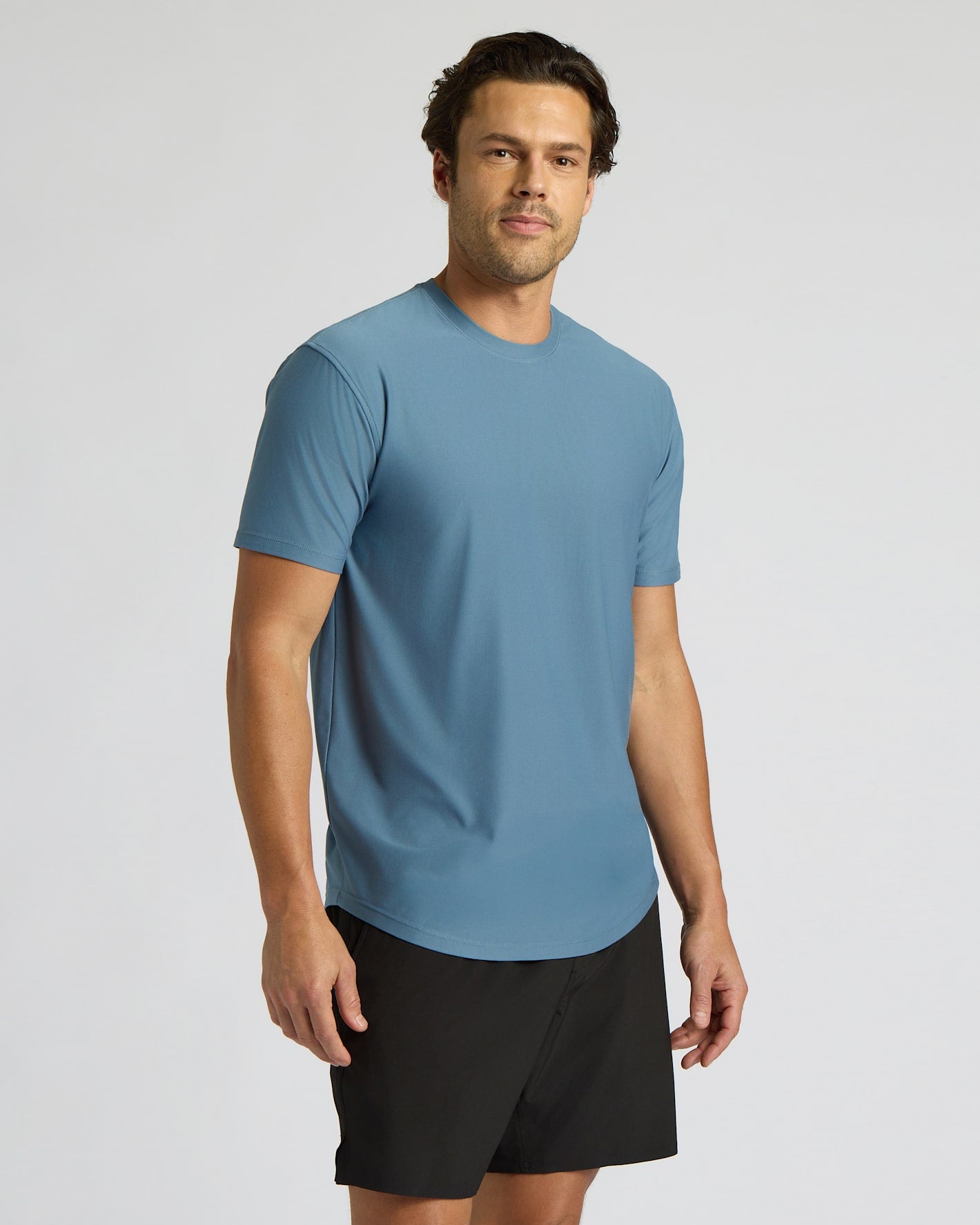 A man wearing a light blue crew-neck athletic t-shirt and black shorts, standing against a light gray studio background.