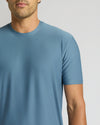 A close-up of a man wearing a short-sleeve crew-neck t-shirt in a dusty slate blue color, shown from the chin down against a light gray studio background.