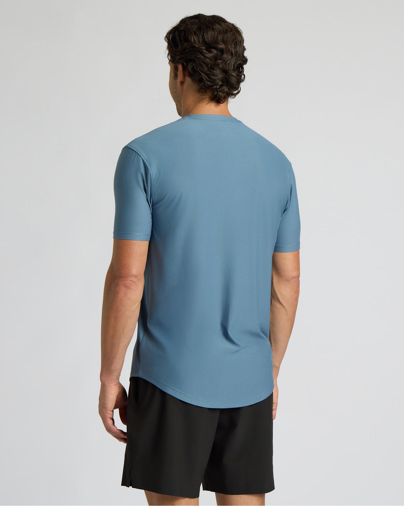 A man viewed from the back wearing a slate blue short-sleeve athletic t-shirt and black shorts against a light gray studio background.