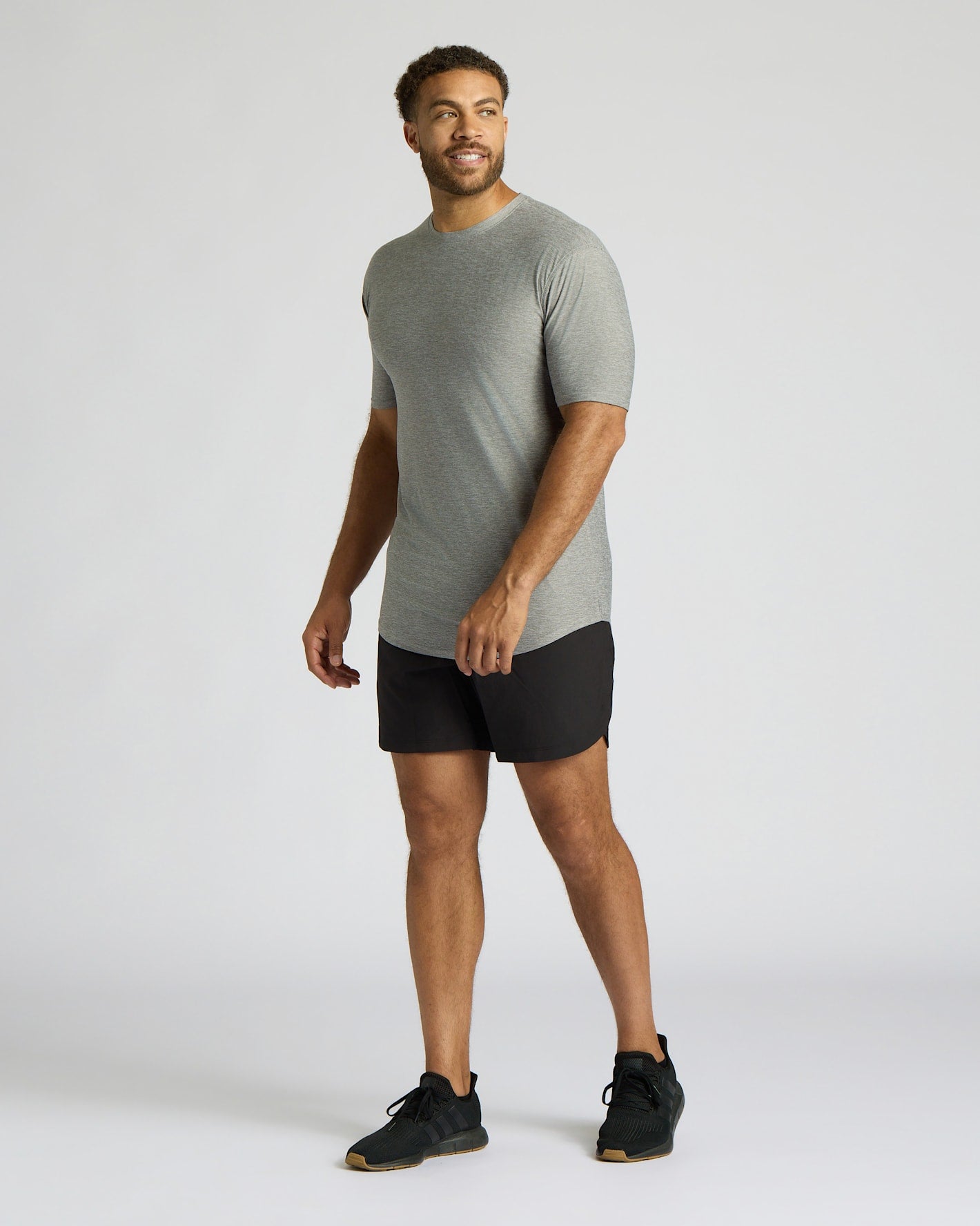 A man modeling a light gray short-sleeve athletic t-shirt and black athletic shorts against a plain light gray studio background.
