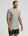 A man wearing a light gray heather crewneck t-shirt and black athletic shorts, standing against a light gray studio background.