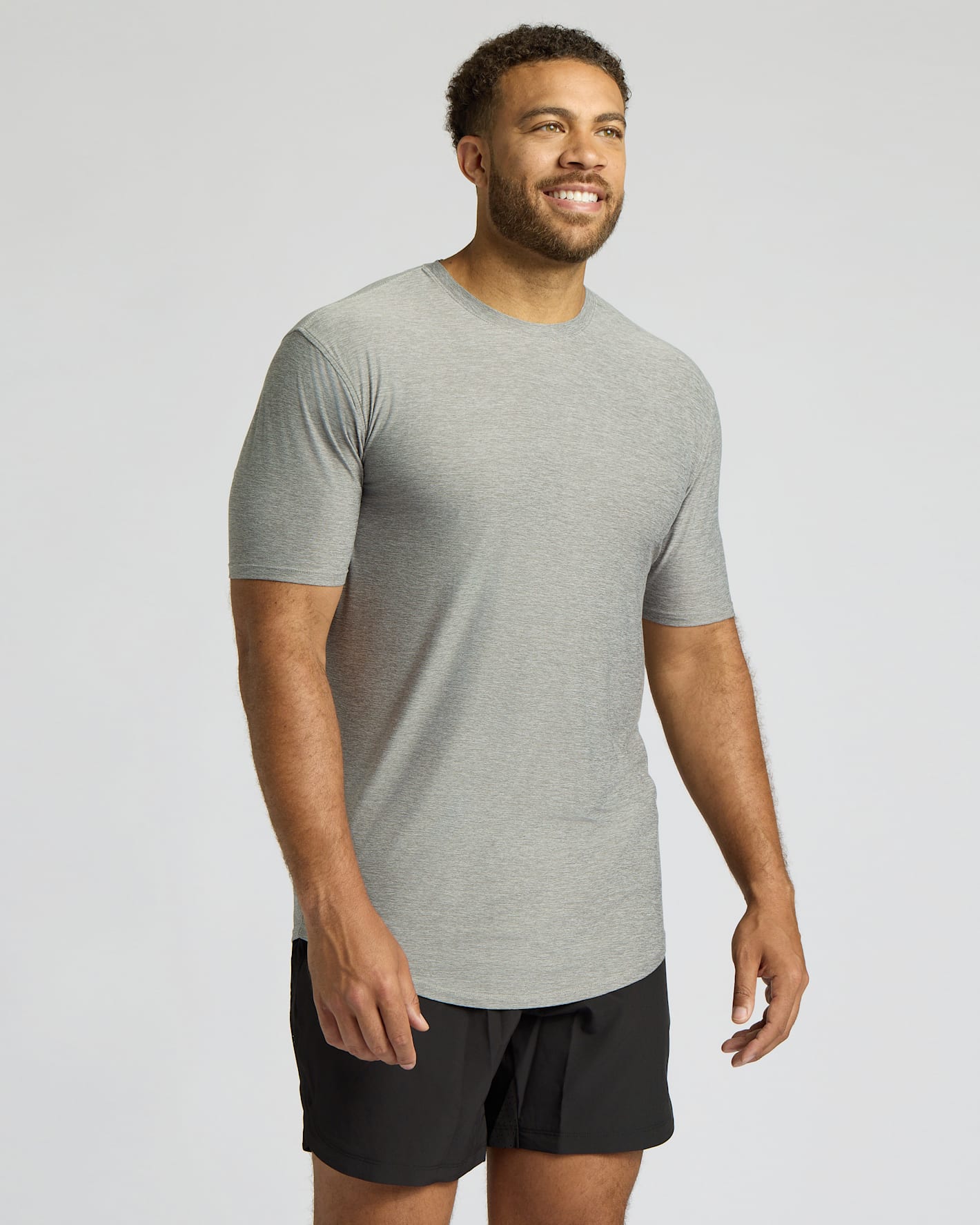 A man wearing a light gray heather crewneck t-shirt and black athletic shorts, standing against a light gray studio background.