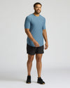A man modeling a short-sleeve athletic t-shirt in slate blue and black athletic shorts, standing against a light gray studio background.