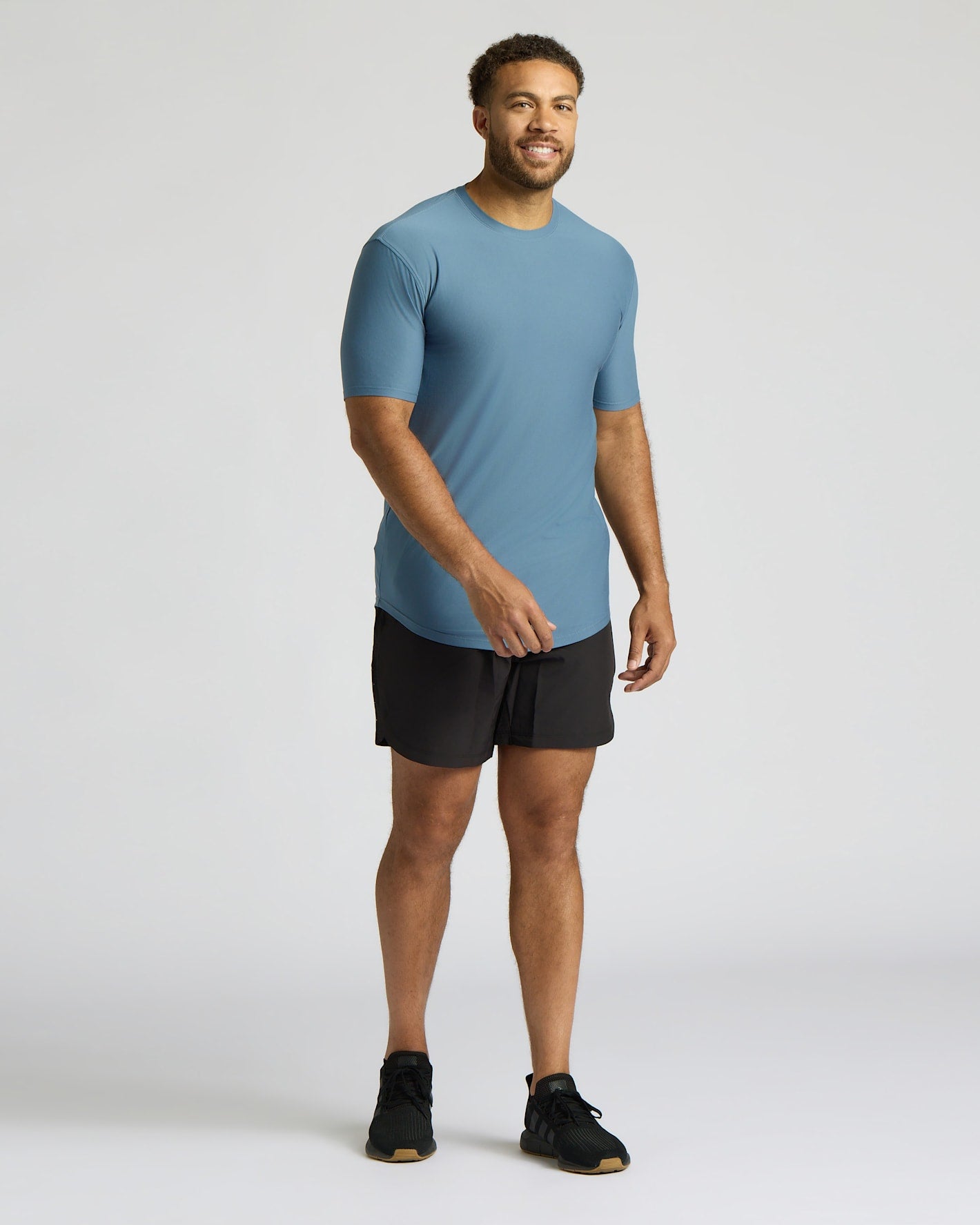 A man modeling a short-sleeve athletic t-shirt in slate blue and black athletic shorts, standing against a light gray studio background.