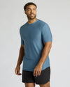 A man wearing a fitted short-sleeve athletic t-shirt in slate blue and black shorts, standing against a light gray studio background.