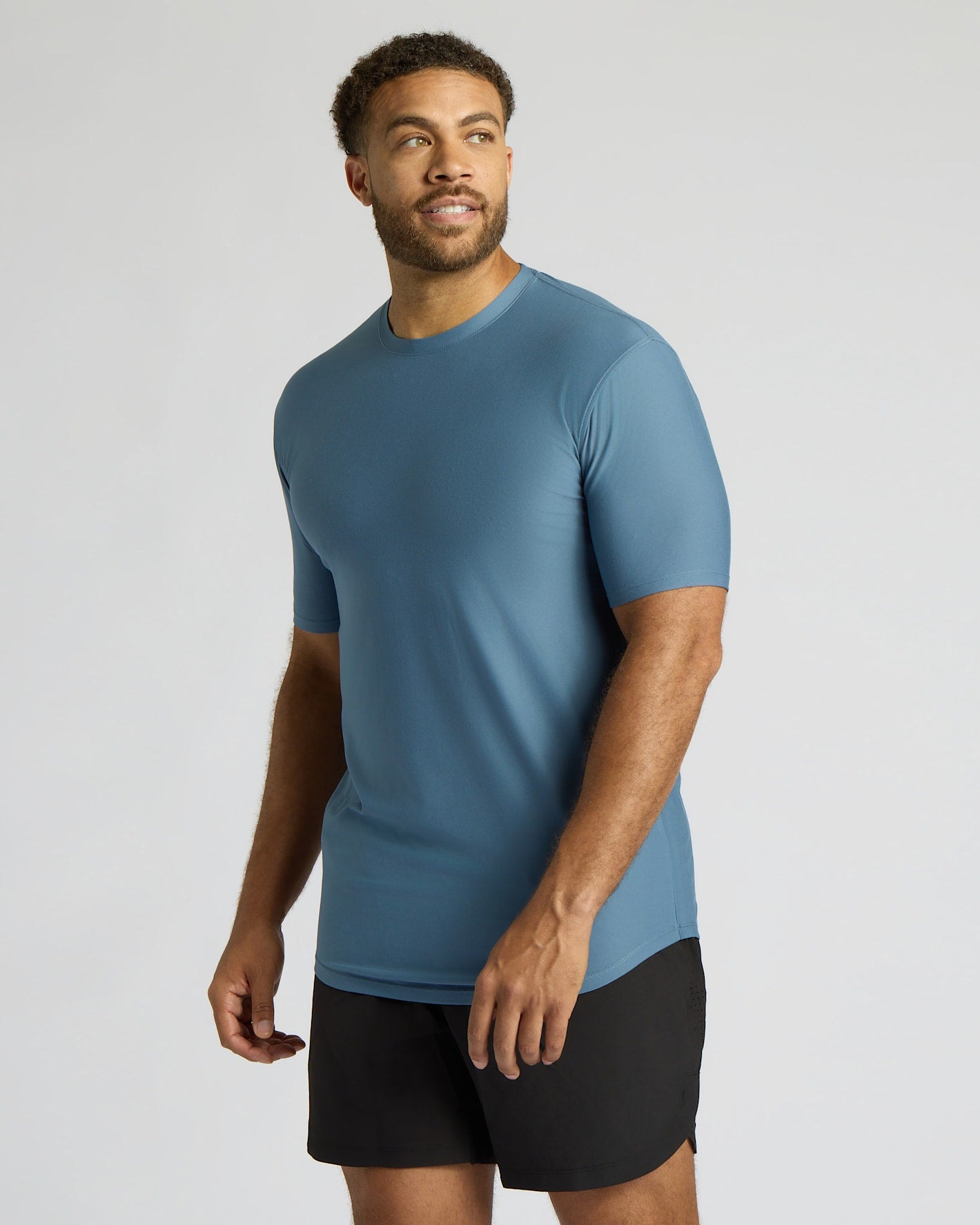 A man wearing a fitted short-sleeve athletic t-shirt in slate blue and black shorts, standing against a light gray studio background.