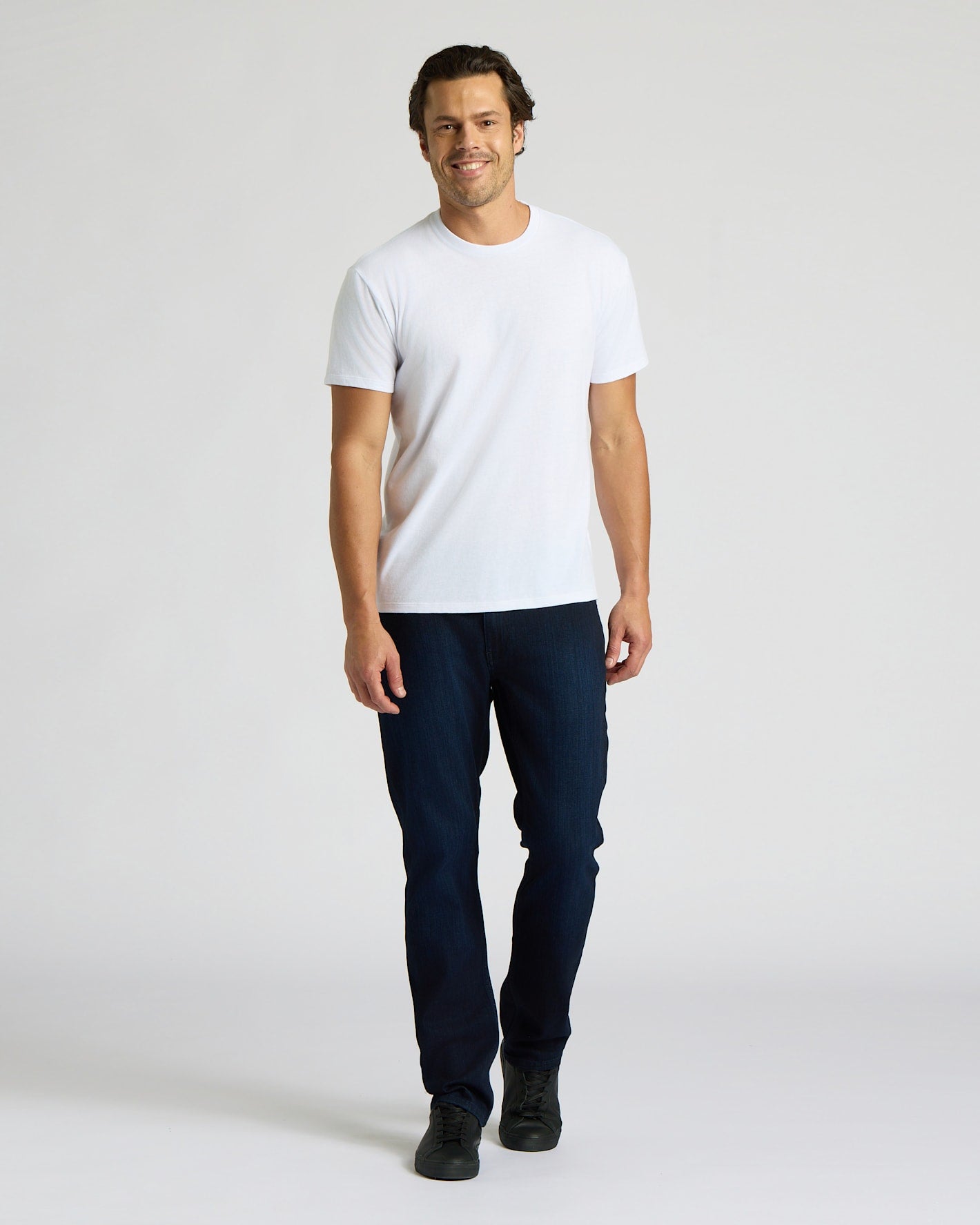 Man wearing a white t-shirt and dark jeans on a plain background. MODEL_INFO: Model is 6'0" wearing size L short