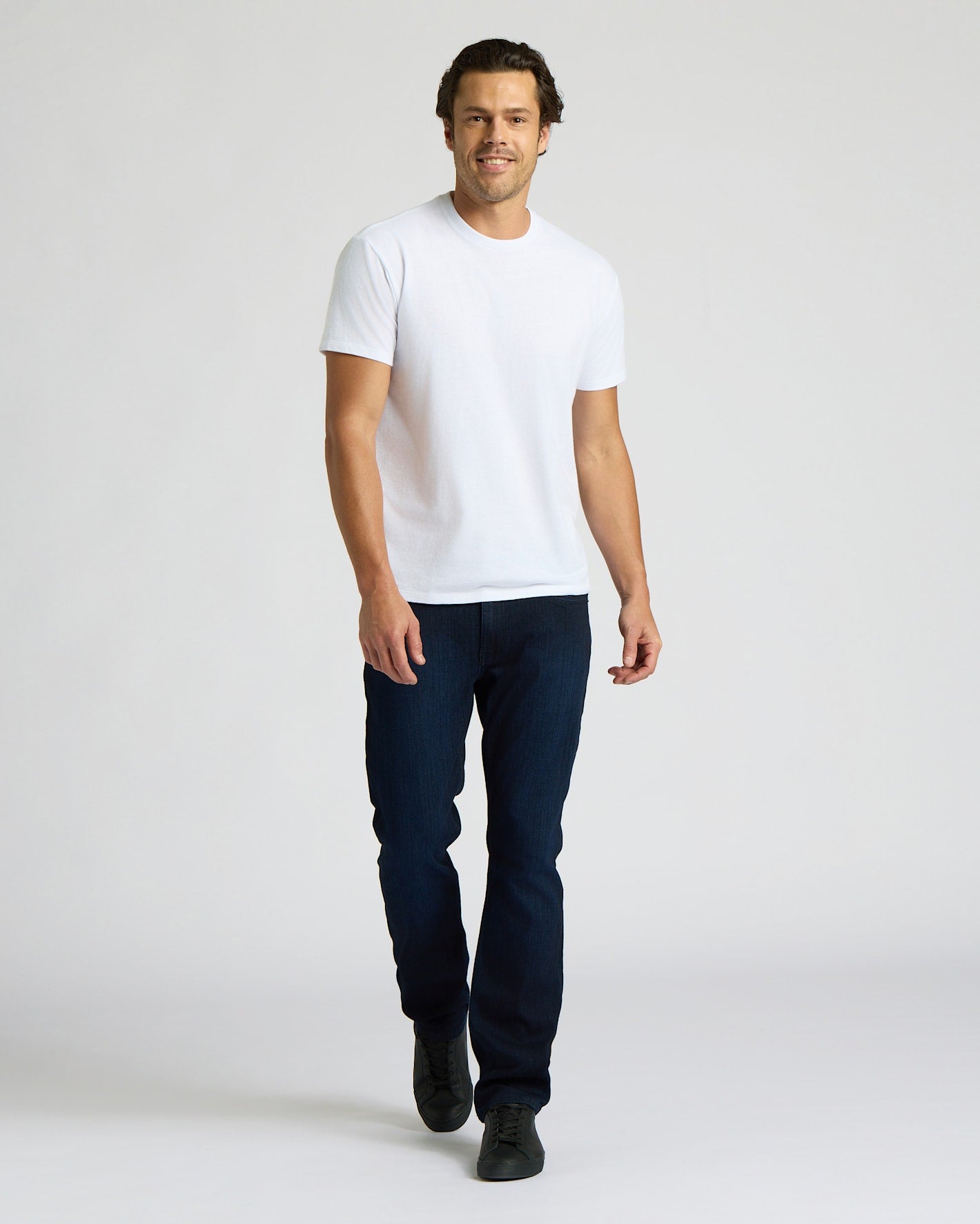 Man wearing a white t-shirt and dark jeans on a plain background. MODEL_INFO: Model is 6'0" wearing size L short