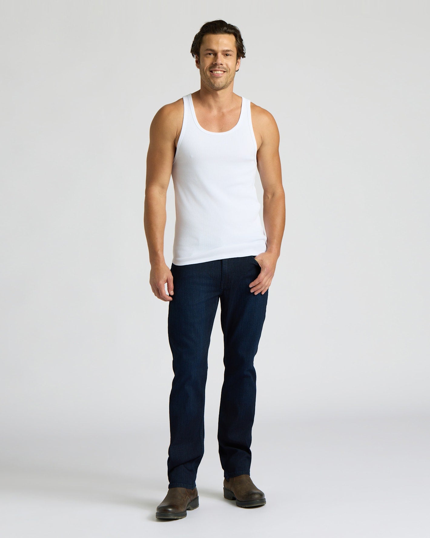 Man wearing a white tank top and dark jeans on a plain background