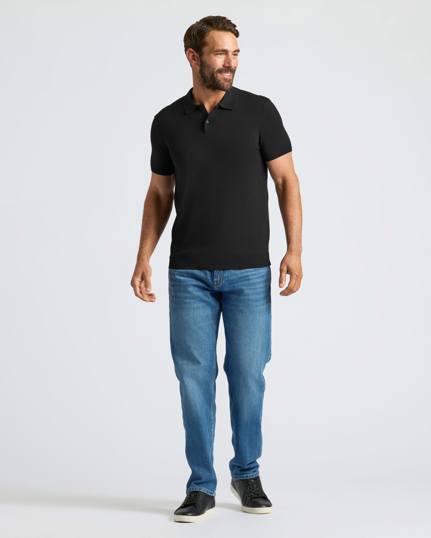 Man wearing Black Dressy Polo, jeans, sneakers, smiling sideways.