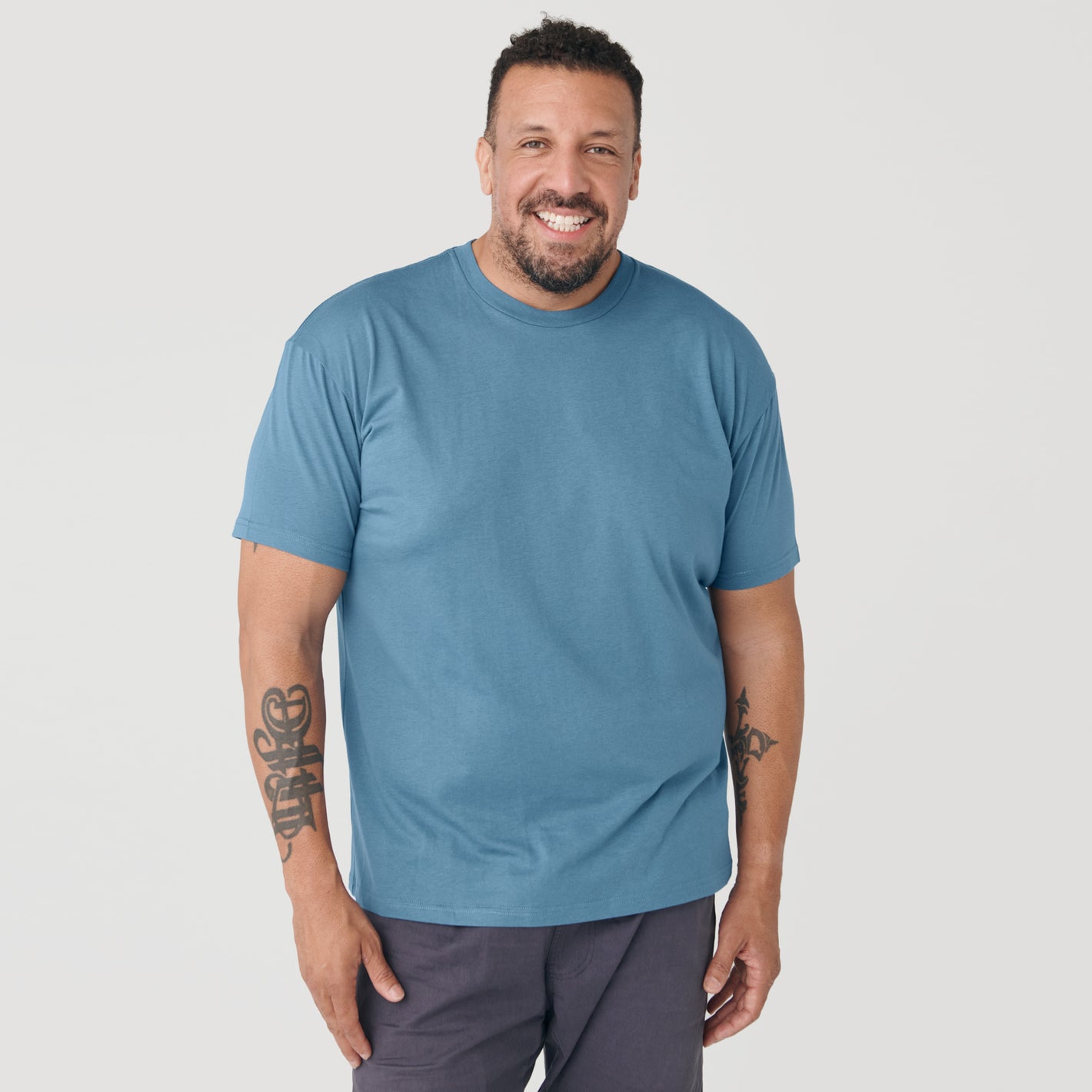 Man wearing Bluestone Short Sleeve Crew Neck Tee, smiling, tattooed forearms.