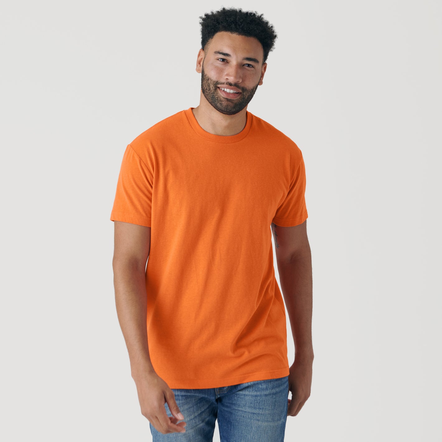 Man wearing Orange Short Sleeve Crew Neck Tee, jeans; smiling slightly.